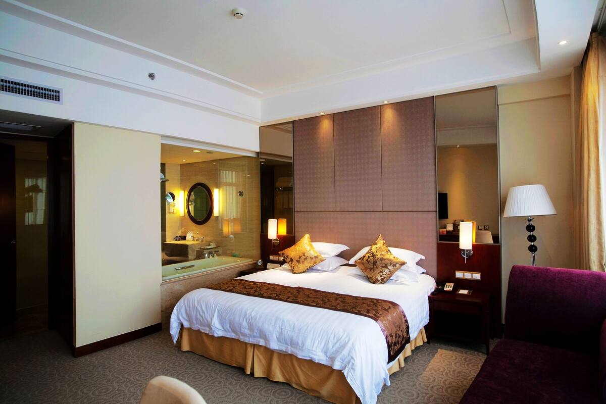 A hotel room with a large bed, a sitting area, a desk, and a bathroom.