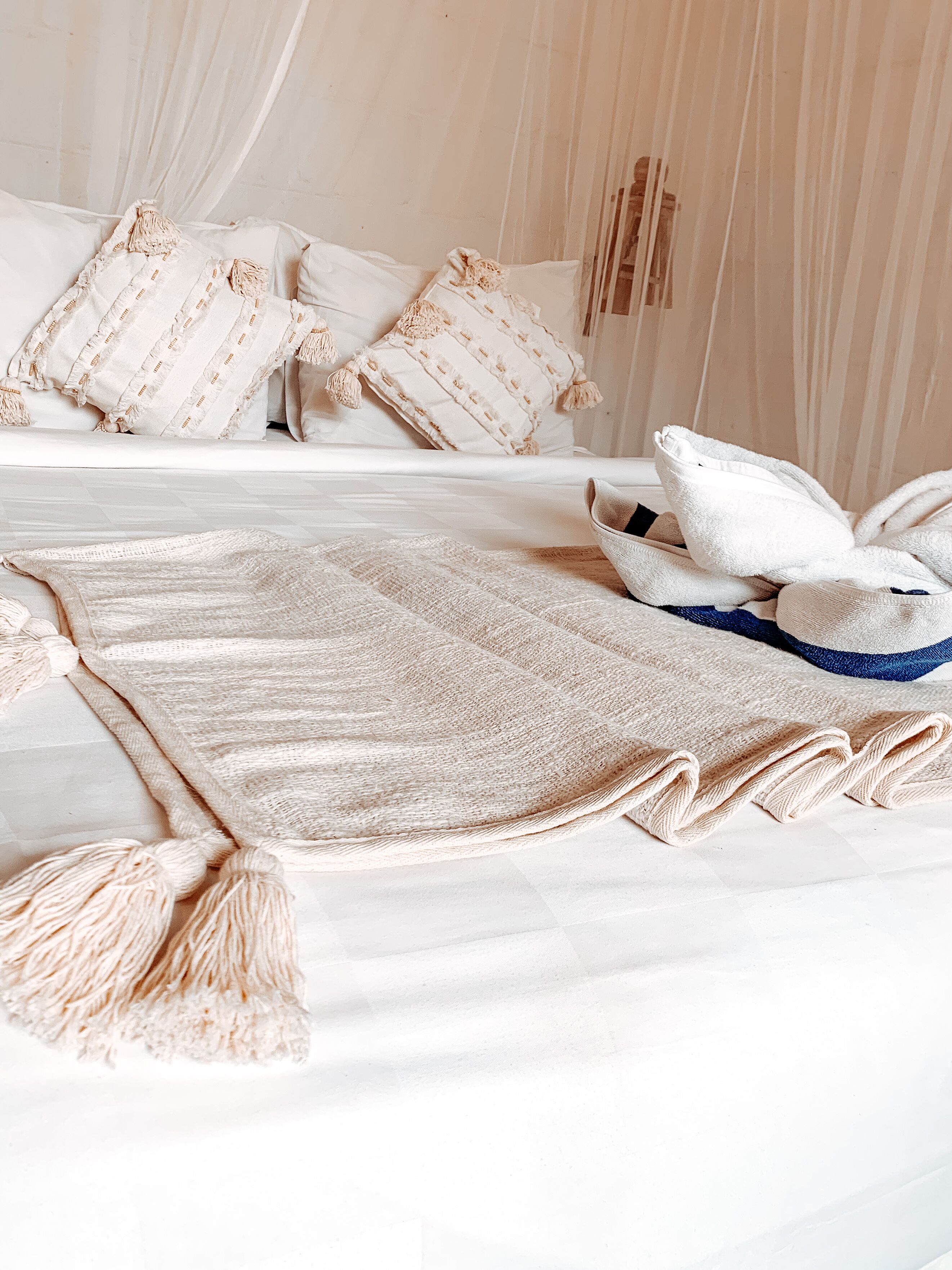 Un lit bien fait, avec une couverture beige et des oreillers, un plaid bleu et beige, et un ciel de lit blanc.