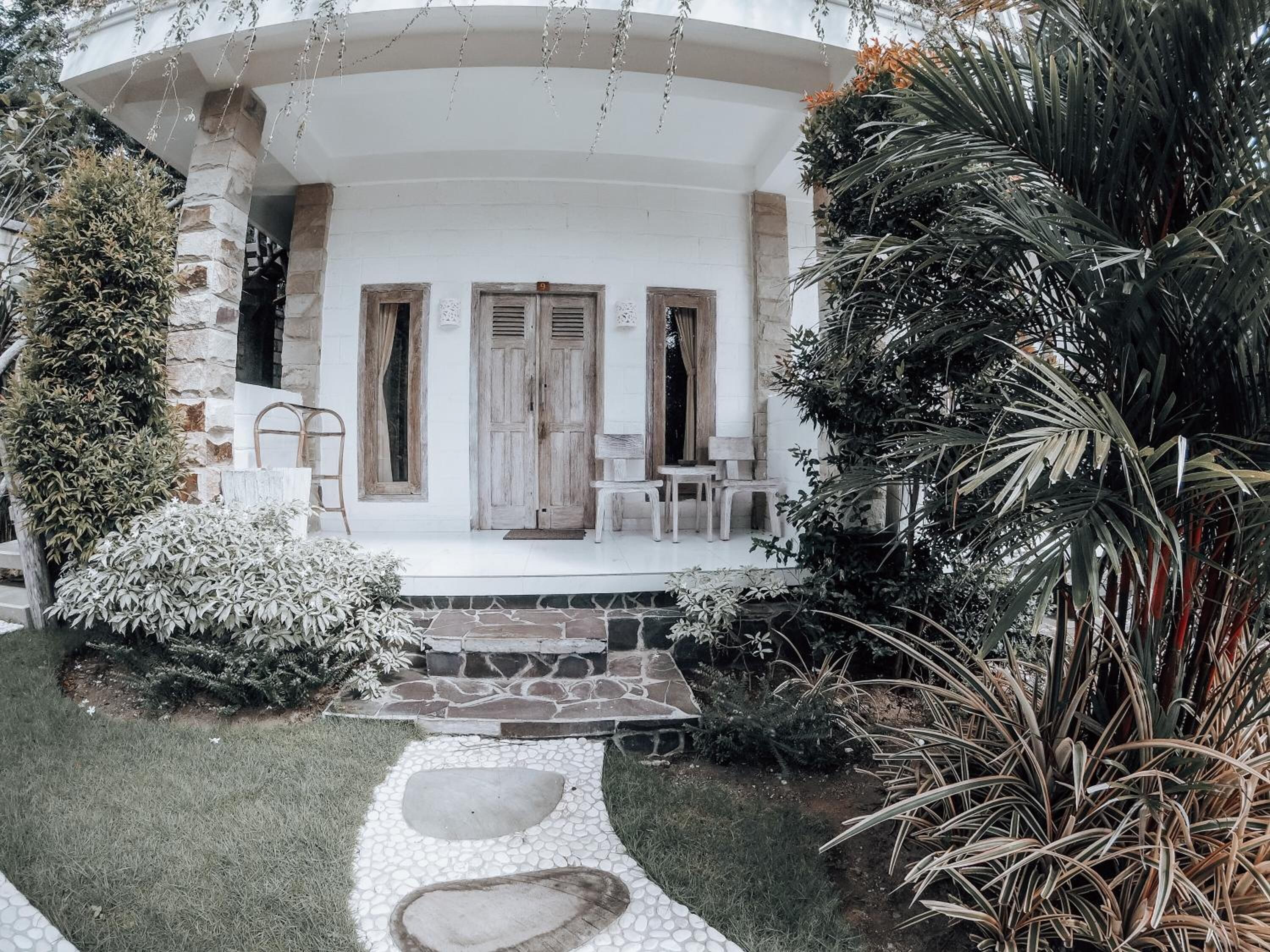 A white building with stone walls, wooden doors, and a stone pathway leading to a seating area with white chairs and tables.