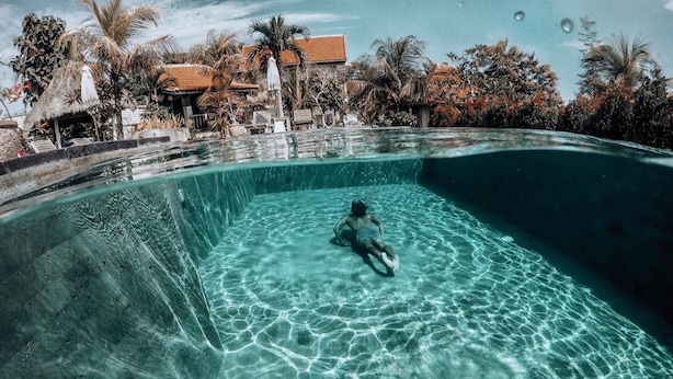 A clear swimming pool with a person swimming, surrounded by palm trees and a building with a tiled roof. - Uluwatu Breeze Village (Pecatu)