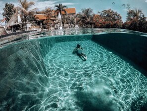 Ein klares Schwimmbecken mit einer Person, die schwimmt, umgeben von Palmen und einem Gebäude mit Ziegeldach.