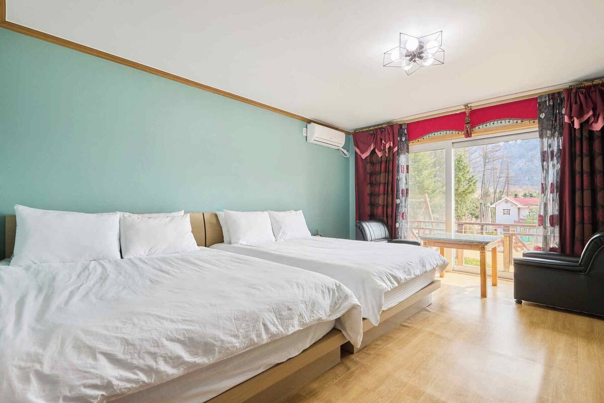 A hotel room with two beds, a small table, and a view of a house through the window.