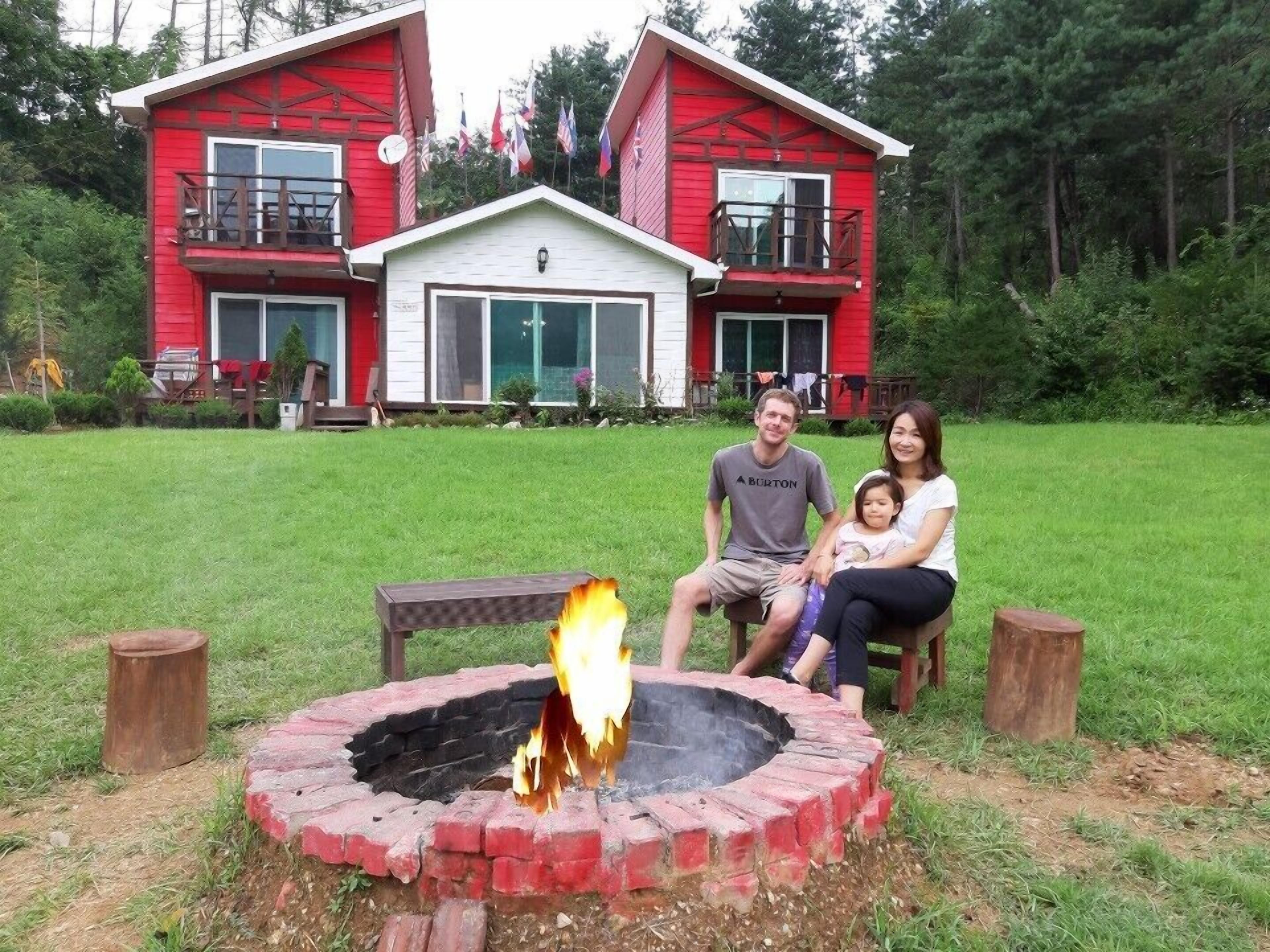 A family sitting by a fire pit in front of a red building with a balcony.