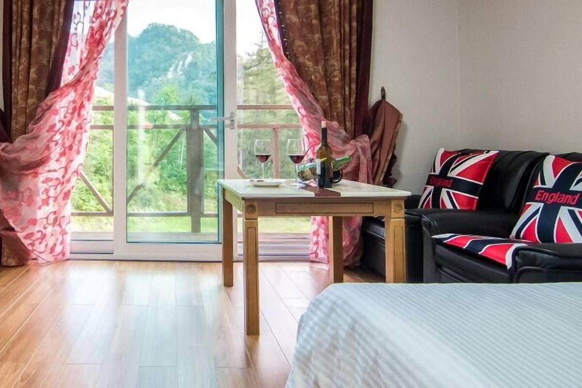 A room with a bed, a desk with a chair, a table with a bottle and glasses, and a view of greenery through the window.