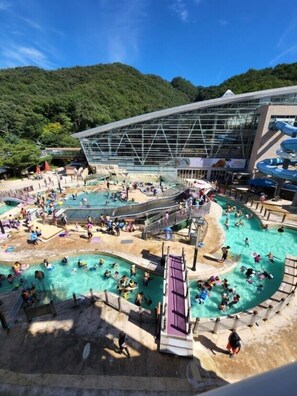 Ein großzügiger Poolbereich im Freien mit mehreren Pools und Wasserrutschen, umgeben von einem modernen Gebäude mit Glasfassade.