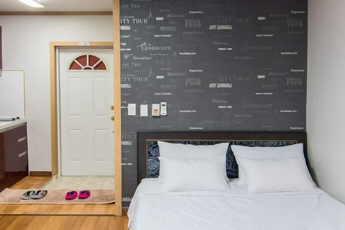 A compact hotel room with a kitchenette, a bed with white bedding, and a wall with travel-related words.