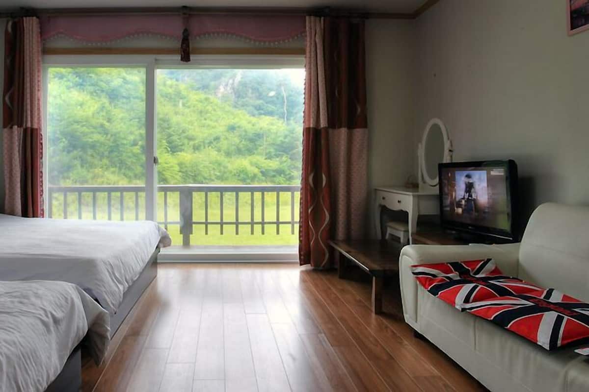 A bedroom with two beds, a television, a sofa, and a balcony with a view of greenery.