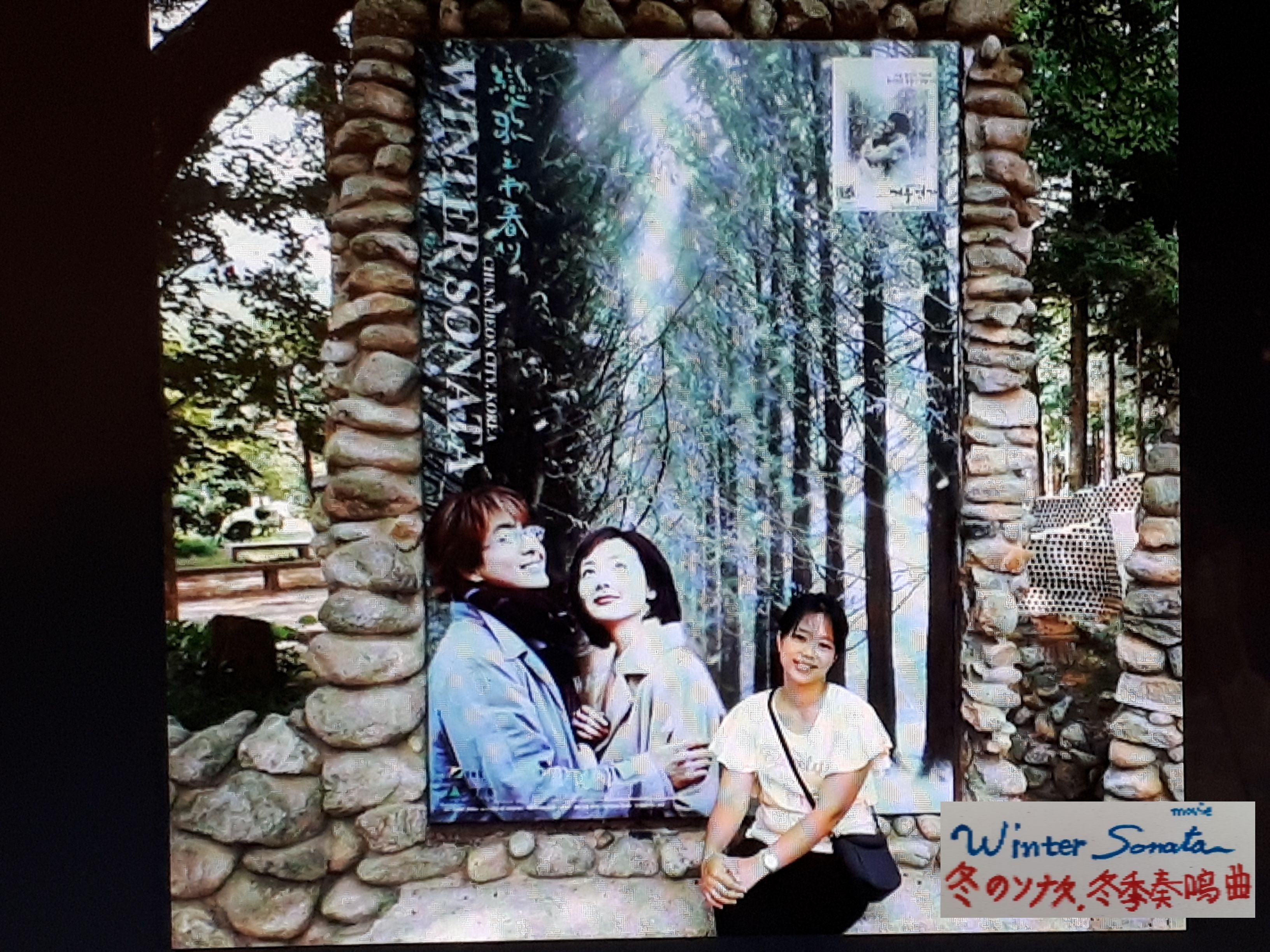 A framed photo of two people in a forest setting, with a stone wall and a sign indicating 'Winter Sonata'.