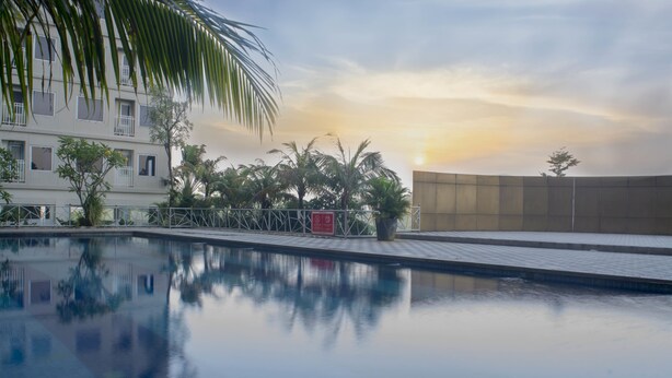 Una piscina con un reflejo cristalino, rodeada por una valla, palmeras y un edificio con balcones.