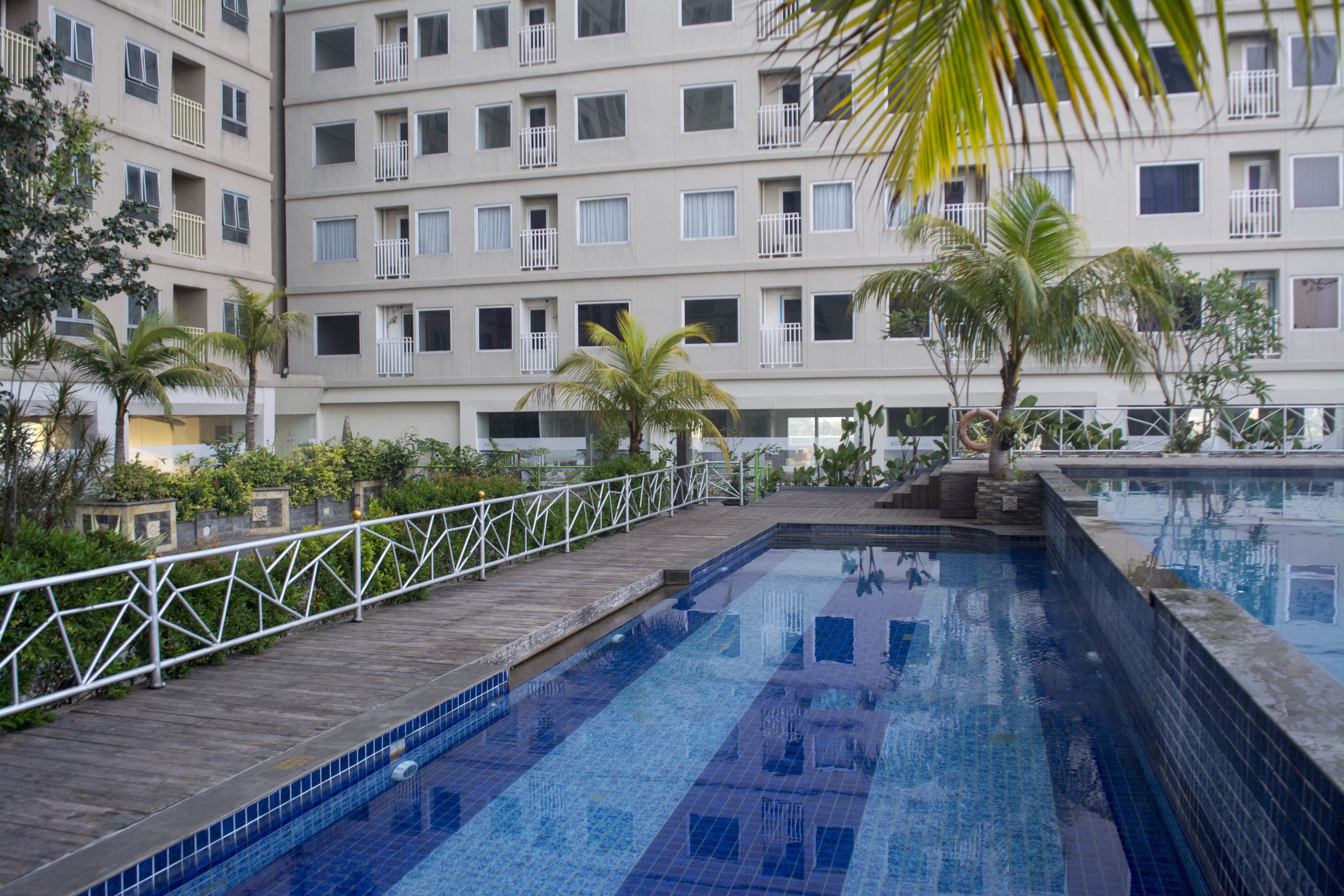 A swimming pool with a wooden deck and metal railing, surrounded by palm trees and a multi-story building with balconies.