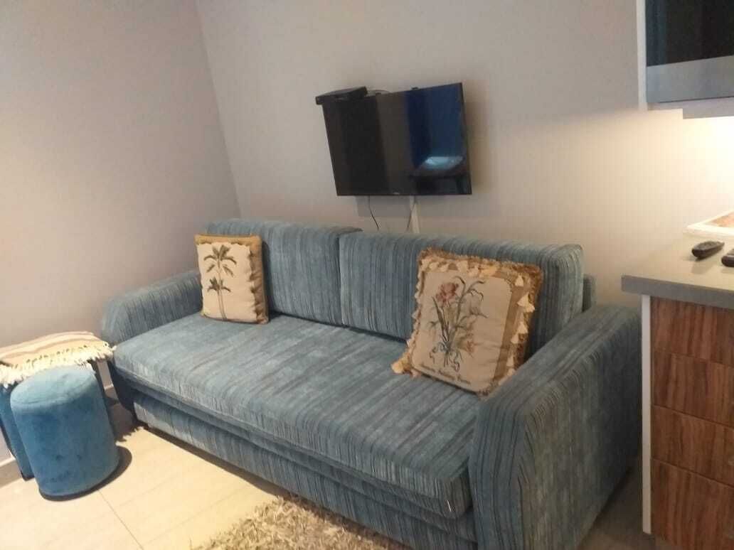 A living room with a blue sofa, two decorative pillows, a small ottoman, and a flat-screen TV mounted on the wall.