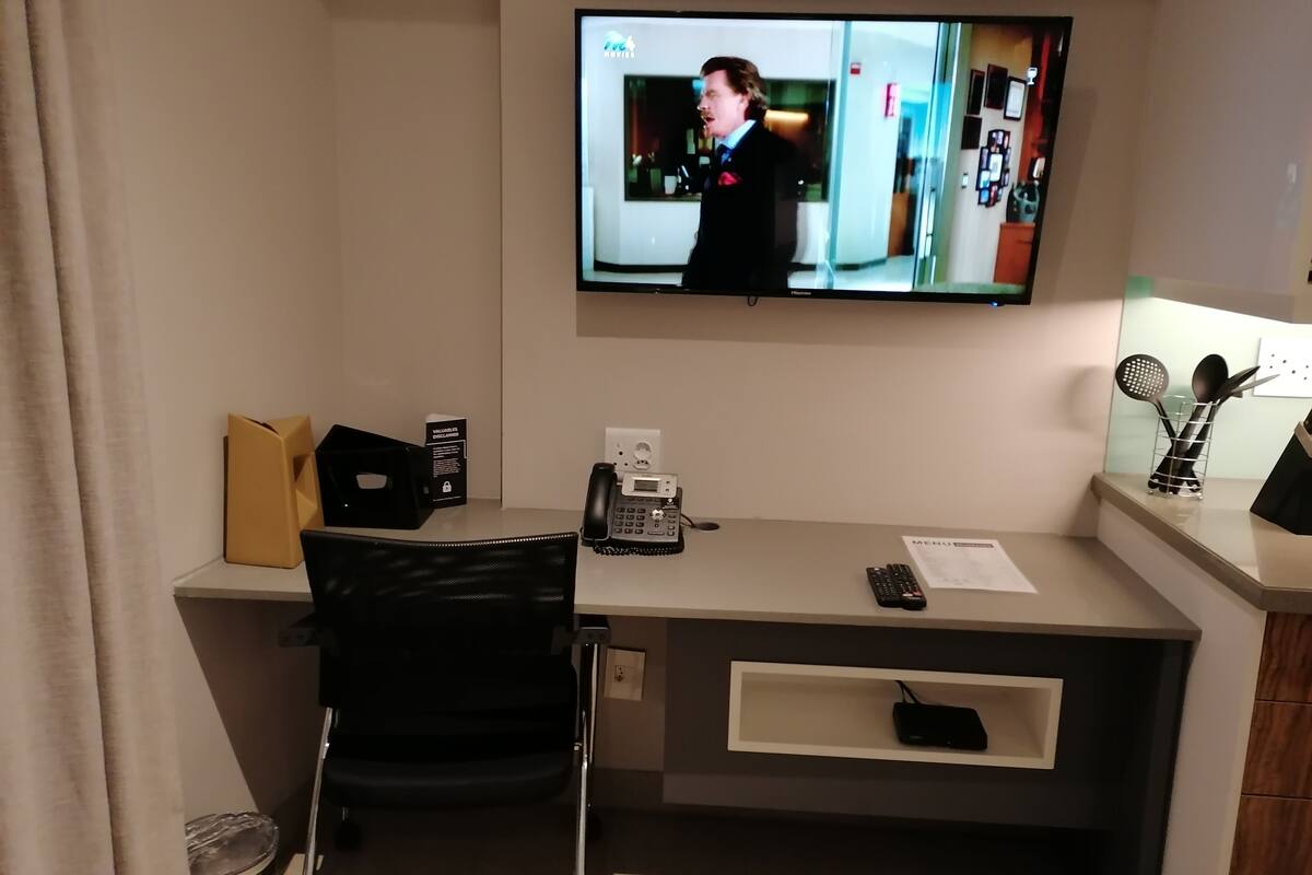 Une chambre d’hôtel équipée d’un bureau, d’une chaise, d’un téléviseur fixé au mur, d’un téléphone, d’une télécommande et d’une petite étagère sur laquelle se trouve un ordinateur portable.