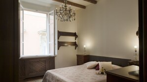 Quarto com uma janela grande, um candelabro, cama com colcha florida e cabeceira de madeira.