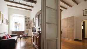 Uma sala de estar com um sofá, um armário de madeira e uma cadeira. Há uma porta que dá para outro quarto.