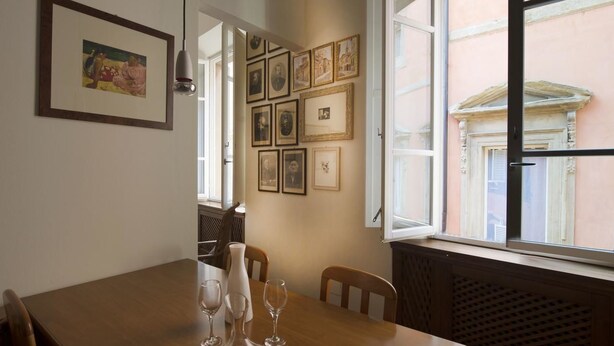 Uma sala de jantar com uma mesa de madeira, cadeiras e um vaso com copos de vinho. Há quadros nas paredes e uma janela com vista para edifícios.