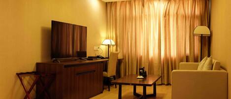 A hotel room with a wooden desk, a television, a lamp, a sofa, and a curtain.