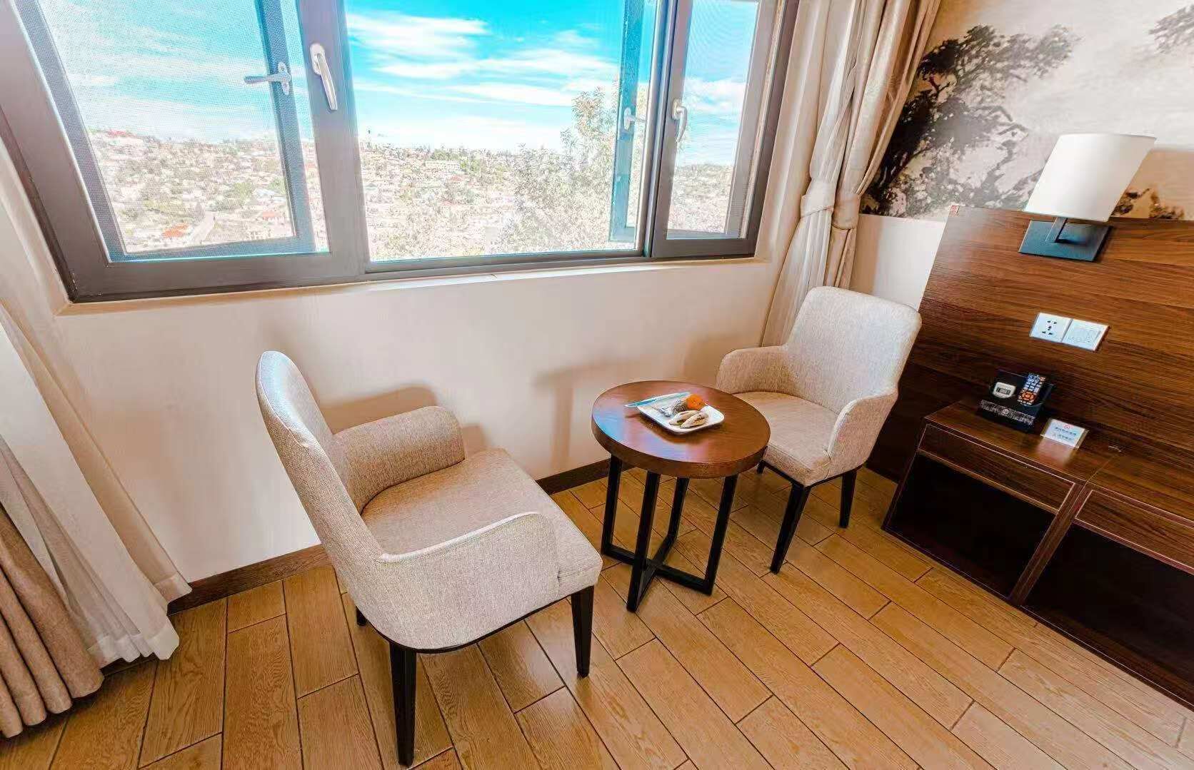 A hotel room with two chairs, a small table, a lamp, a wooden side table, and a view of the city through large windows.