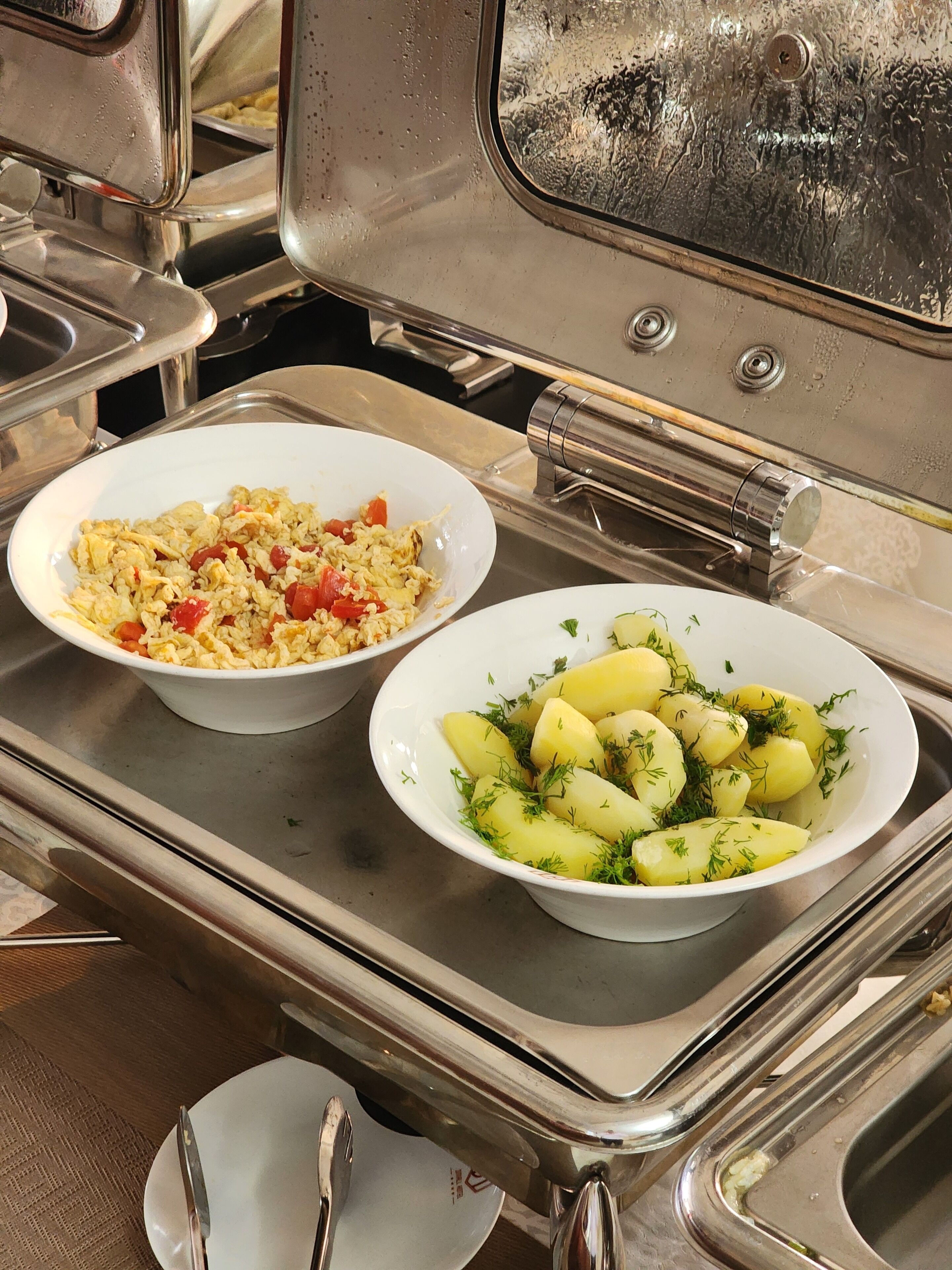 A buffet setup with two bowls of food: one with scrambled eggs and vegetables, and another with boiled potatoes garnished with herbs.