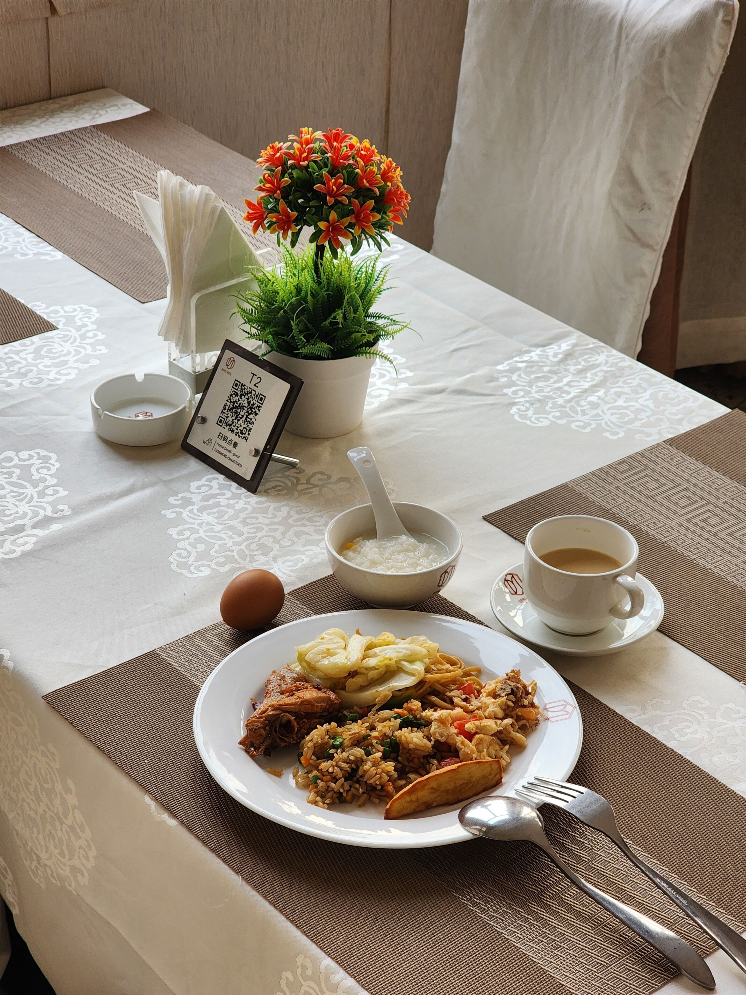 Una mesa de comedor con un plato de comida, una taza de café, un tazón de sopa, una cuchara, un tenedor, un letrero con un código QR, una pequeña planta en maceta y un jarrón decorativo.