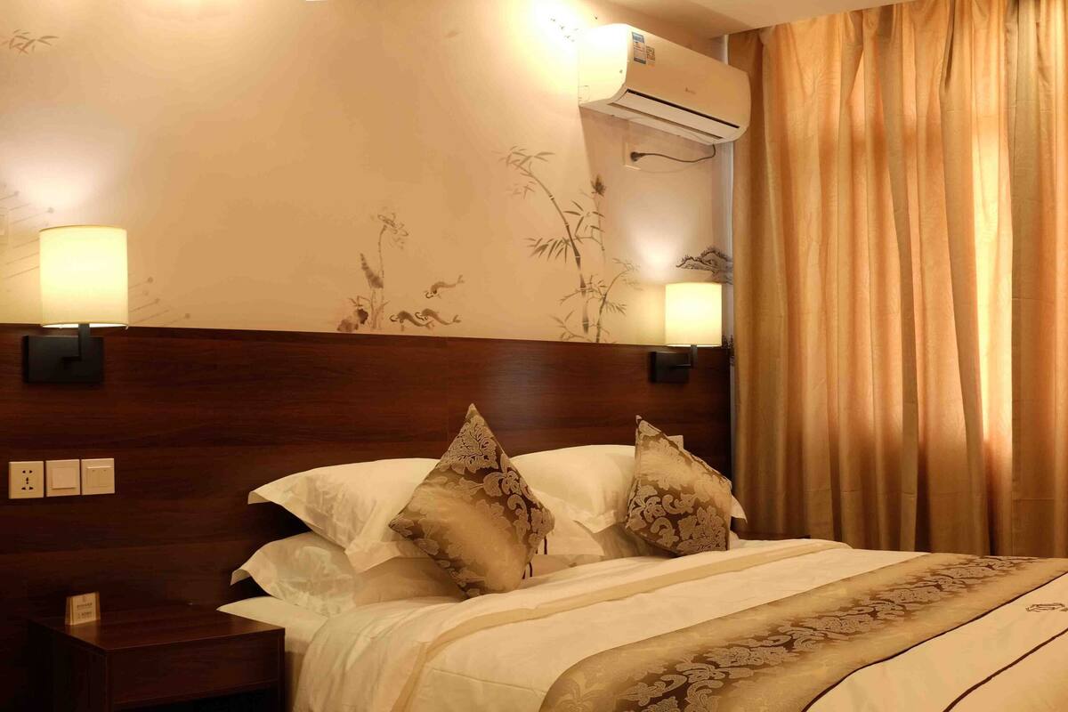 A hotel room with a wooden headboard, a bed with patterned pillows, wall-mounted lights, and a large window with curtains.