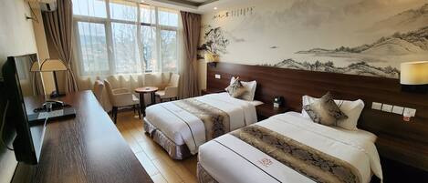 A hotel room with two beds, a desk, a chair, a table, a lamp, and a large wall painting.