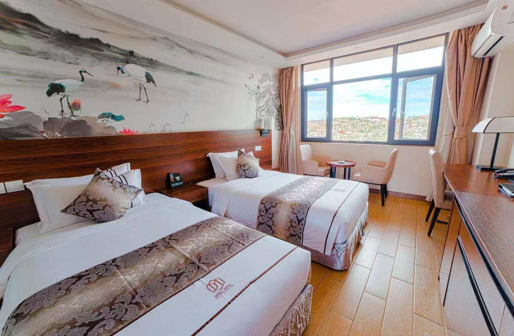 A hotel room with two beds, a desk, a chair, a small table, a painting on the wall, and a large window.
