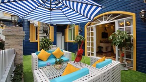 Un espace patio agrémenté d’un parasol à rayures bleues et blanches, d’un canapé en osier blanc avec des coussins jaunes et d’une sculpture rouge.