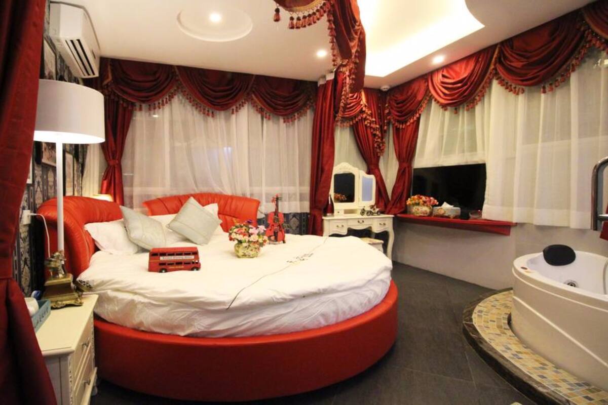 A bedroom with a red circular bed, white bedding, a vanity with a mirror, and a bathtub.