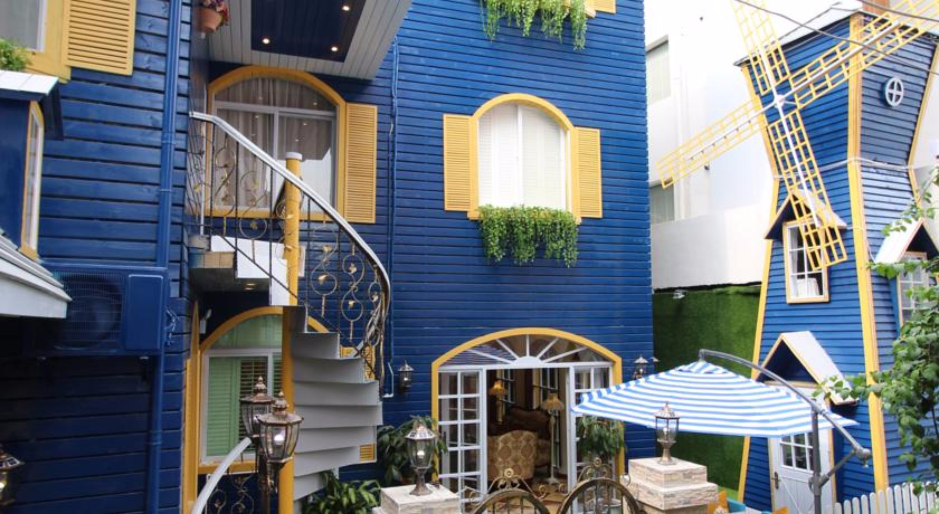 A blue building with a staircase, a window with plants, and a small outdoor seating area with a striped umbrella.