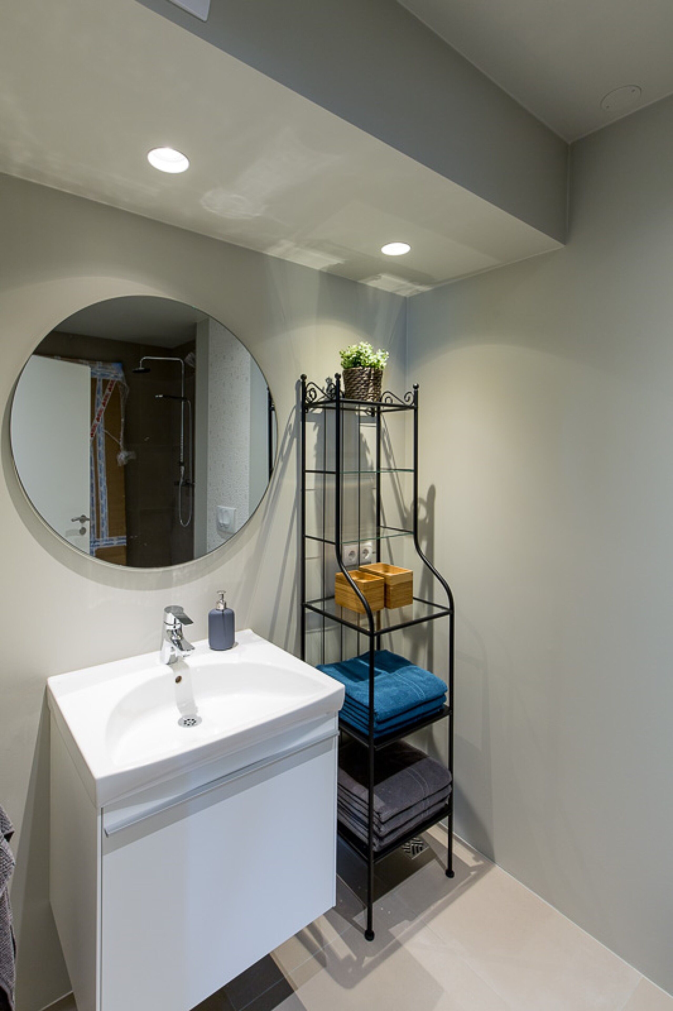 A bathroom with a white sink, a round mirror, and a metal shelf with folded towels.