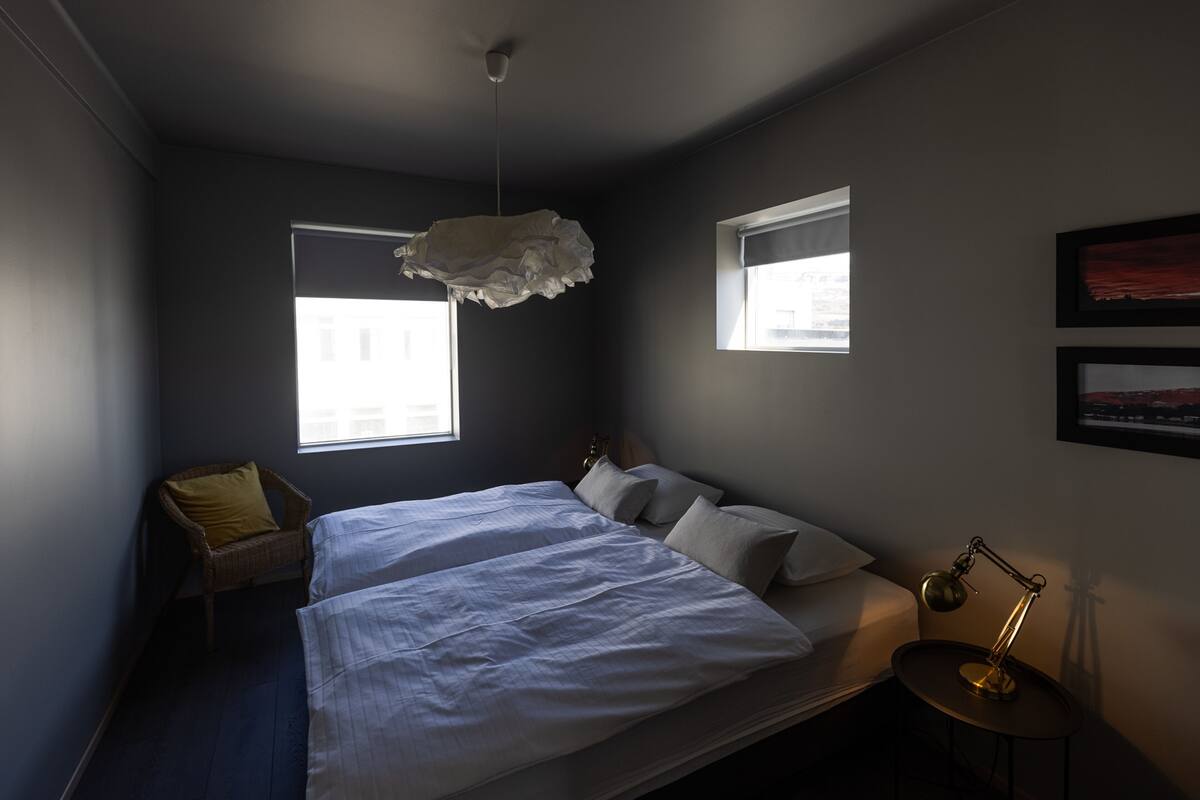 A bedroom with two beds, a hanging light fixture, a window, and a desk with a lamp.