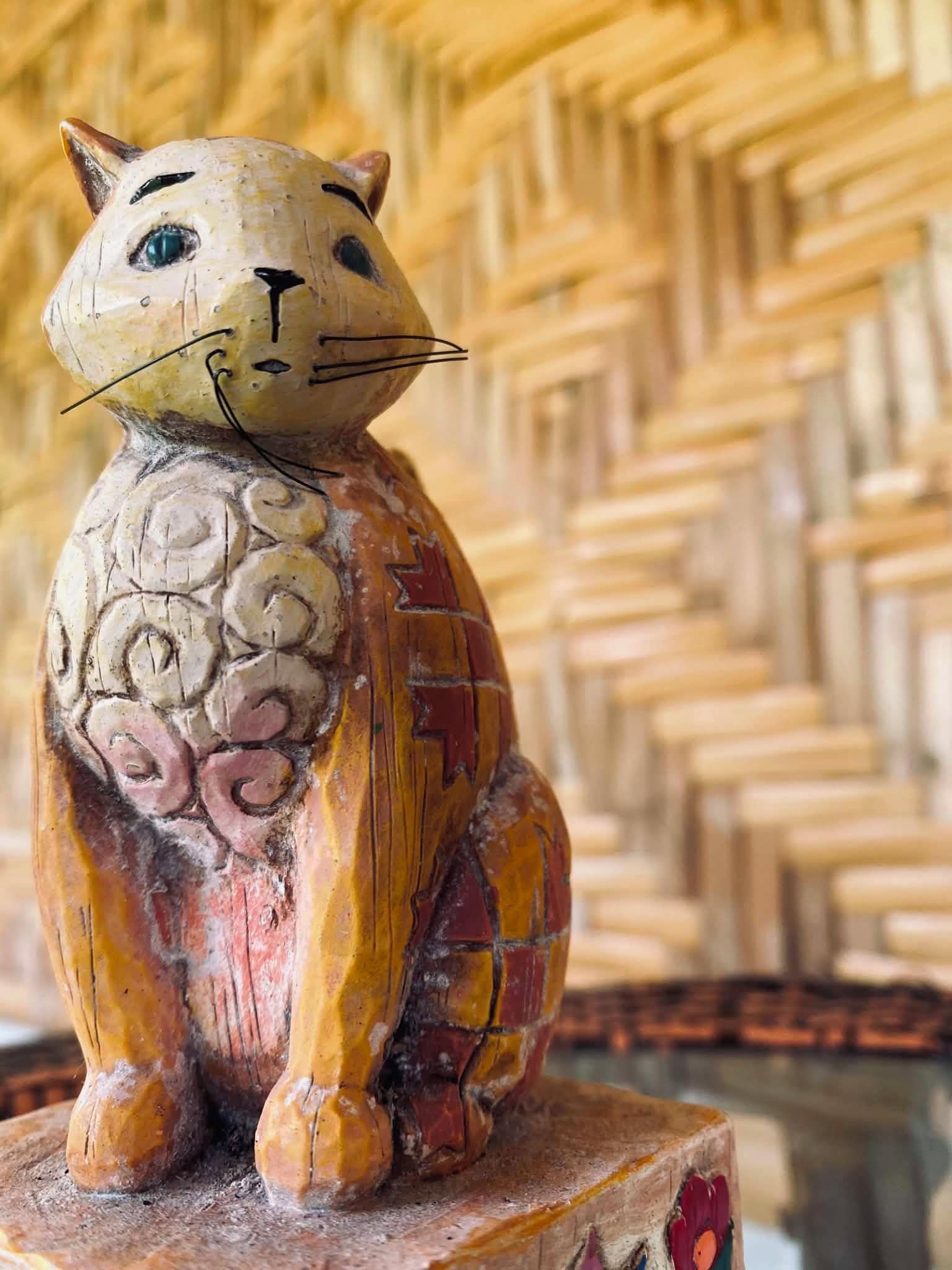Una escultura de madera de un gato con cuerpo texturizado y cara plana, sobre un pedestal de madera.