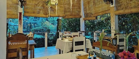 An indoor dining area with tables and chairs, a large window with a view of greenery, and hanging light fixtures.