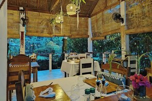 An indoor dining area with tables and chairs, a large window with a view of greenery, and hanging light fixtures. - Nypa Style Resort Camiguin (Mambajao)