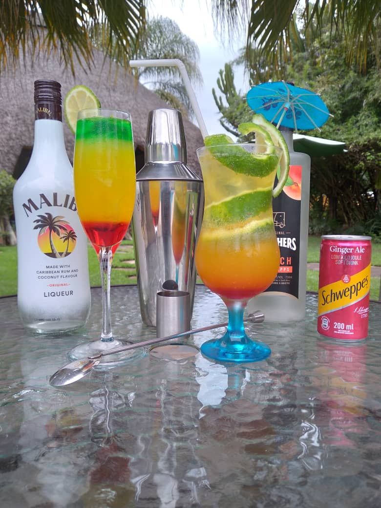 A table with two cocktails, a bottle of Malibu rum, a shaker, a can of Schweppes ginger ale, and a blue umbrella.