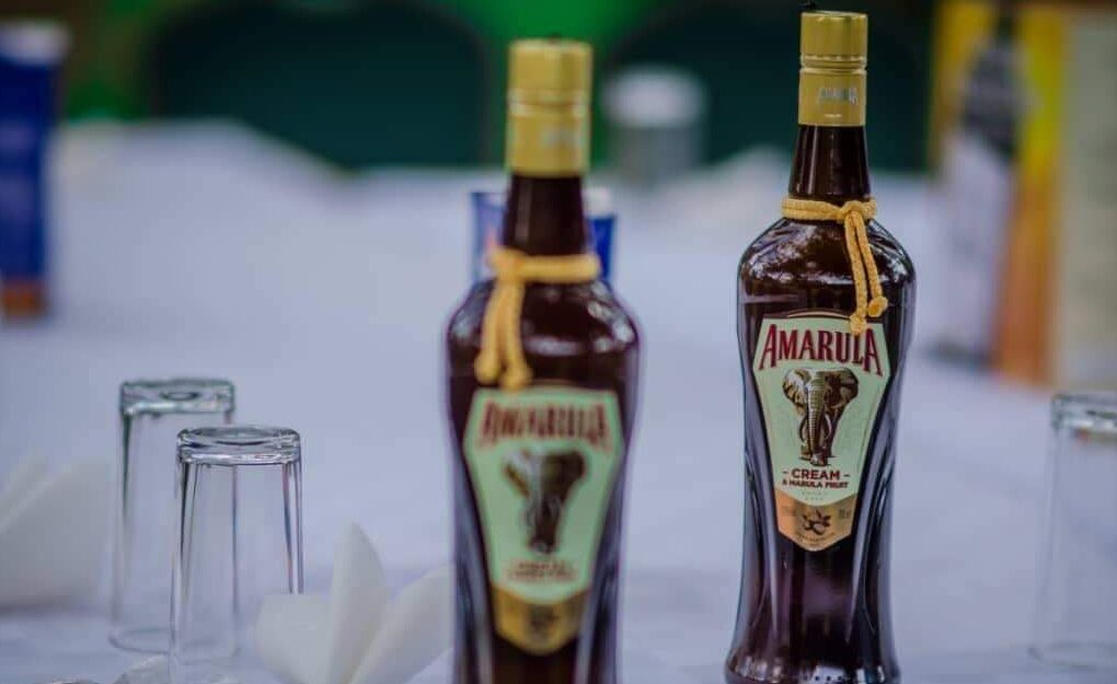 Dos botellas de licor cremoso Amarula con tapa dorada, atadas con una cinta, y dos vasos de cristal sobre una superficie blanca.