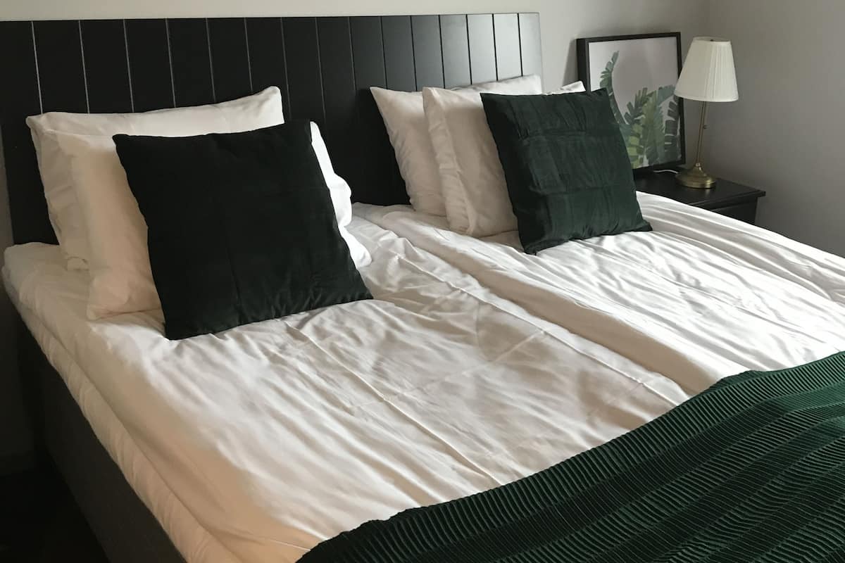 Cama bem arrumada com roupa de cama branca e verde escuro, cabeceira preta e abajur.