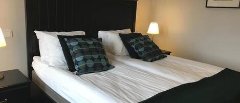 Una cama bien tendida con sábanas blancas y dos almohadones decorativos, un cabecero oscuro y lámparas de noche a cada lado.
