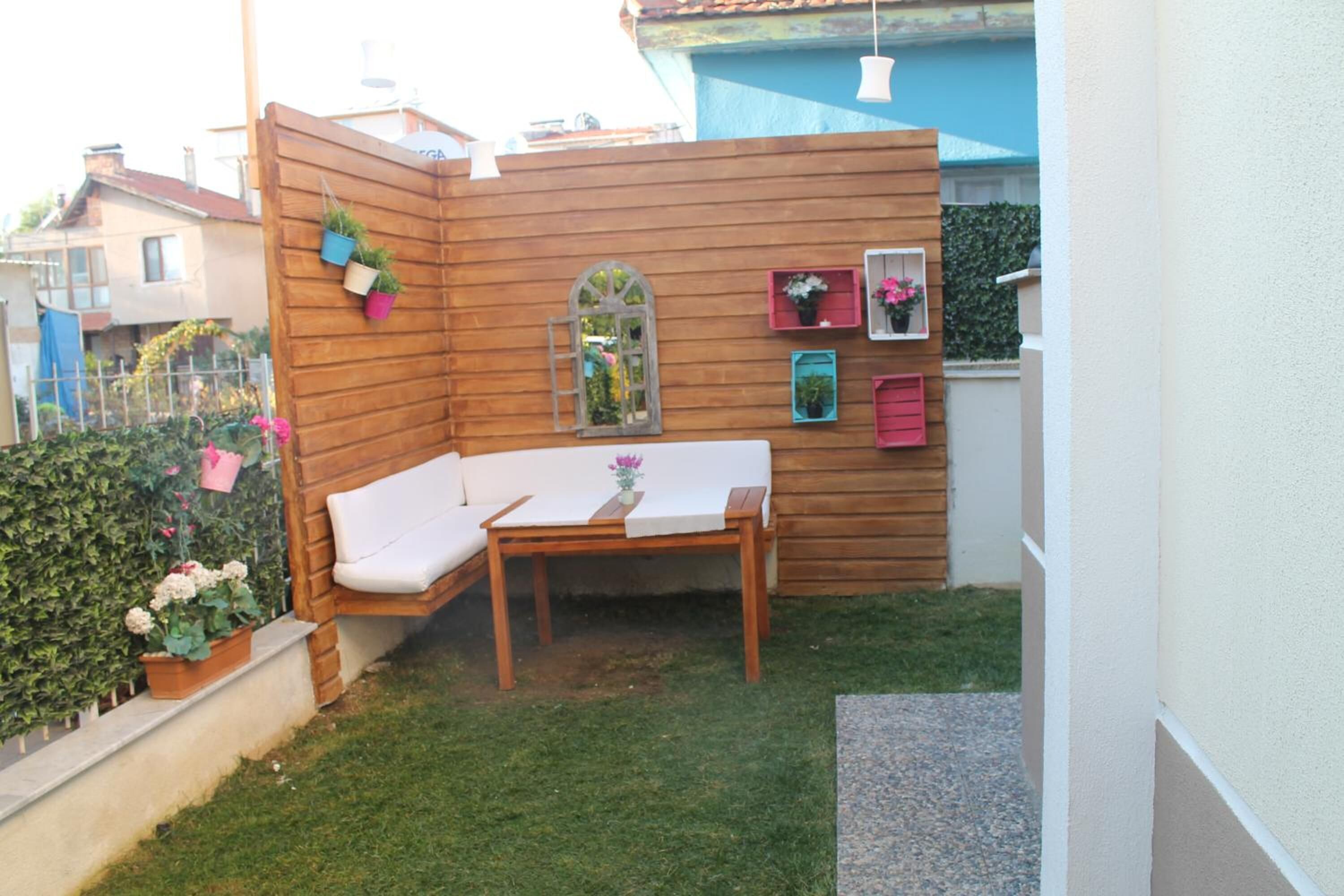 A small outdoor seating area with a wooden bench and table, surrounded by a wooden partition with potted plants and a small window.
