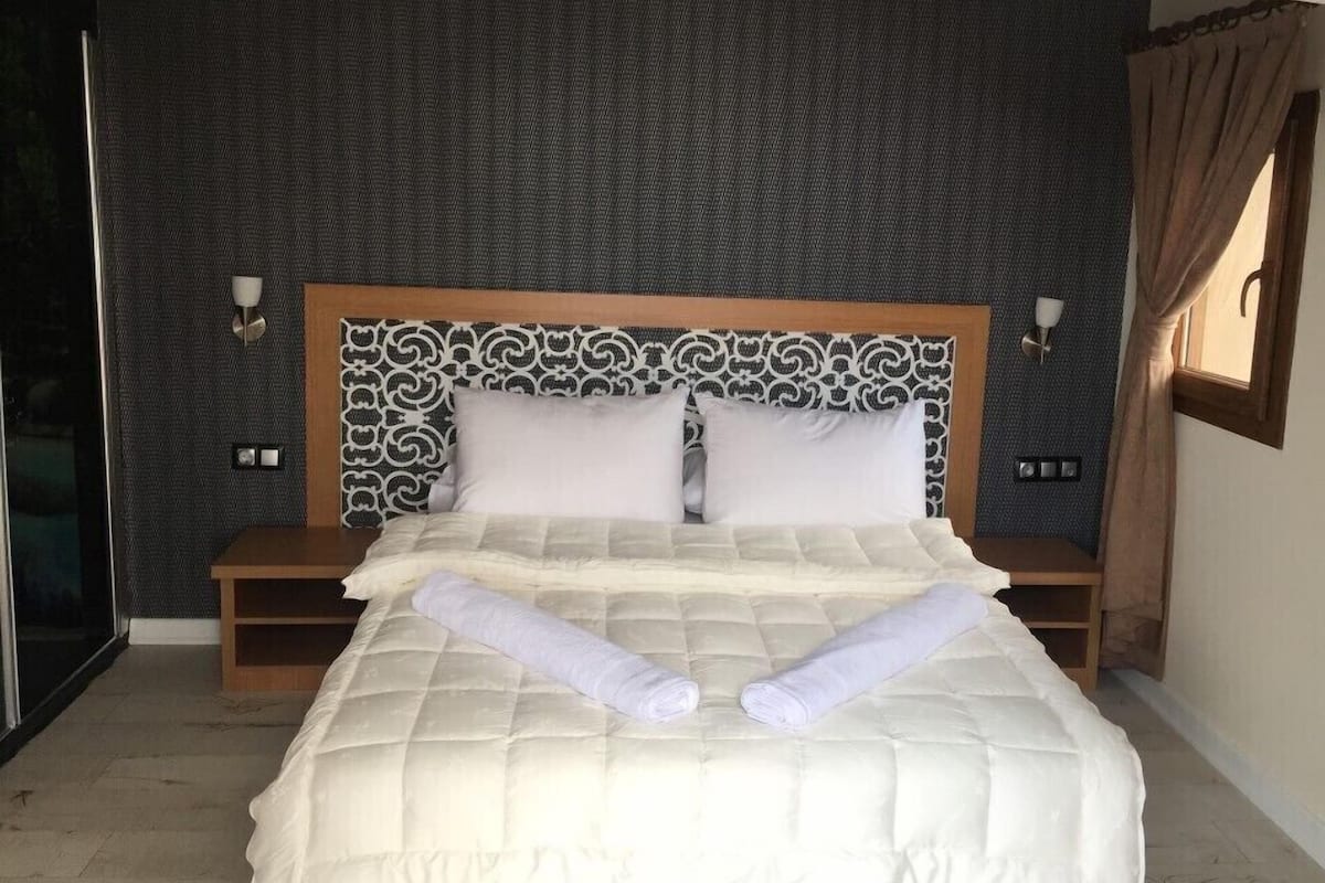 Una cama bien hecha con edredón blanco y dos toallas enrolladas, cabecera de madera con diseño estampado, mesitas de noche con lámparas y una pared oscura con textura.