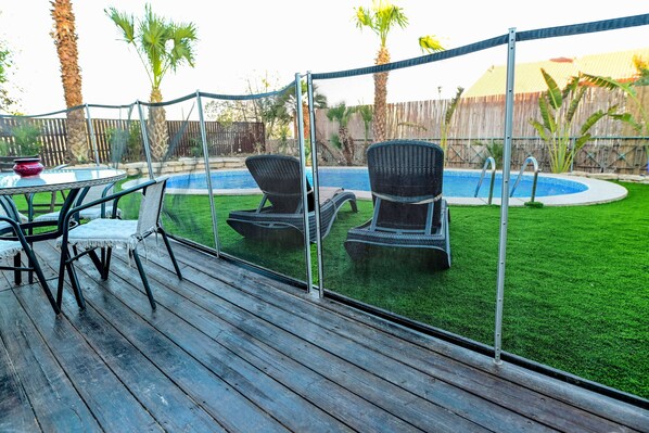 A wooden deck with outdoor furniture, a pool, and a fenced area with palm trees.