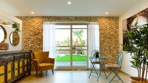 A room with a stone wall, a yellow armchair, a dining table with chairs, and a large window with a view of a garden. - Villa Yotam (Eilat)
