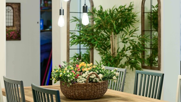 A dining table with a wicker basket filled with flowers, surrounded by chairs, and a hanging light fixture above. - Villa Yotam (Eilat)