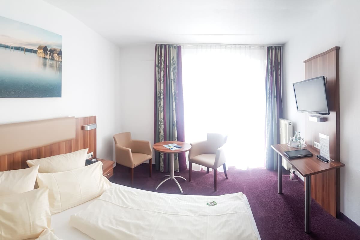 A hotel room with a large bed, a desk with a computer, a chair, a small table, and a TV.