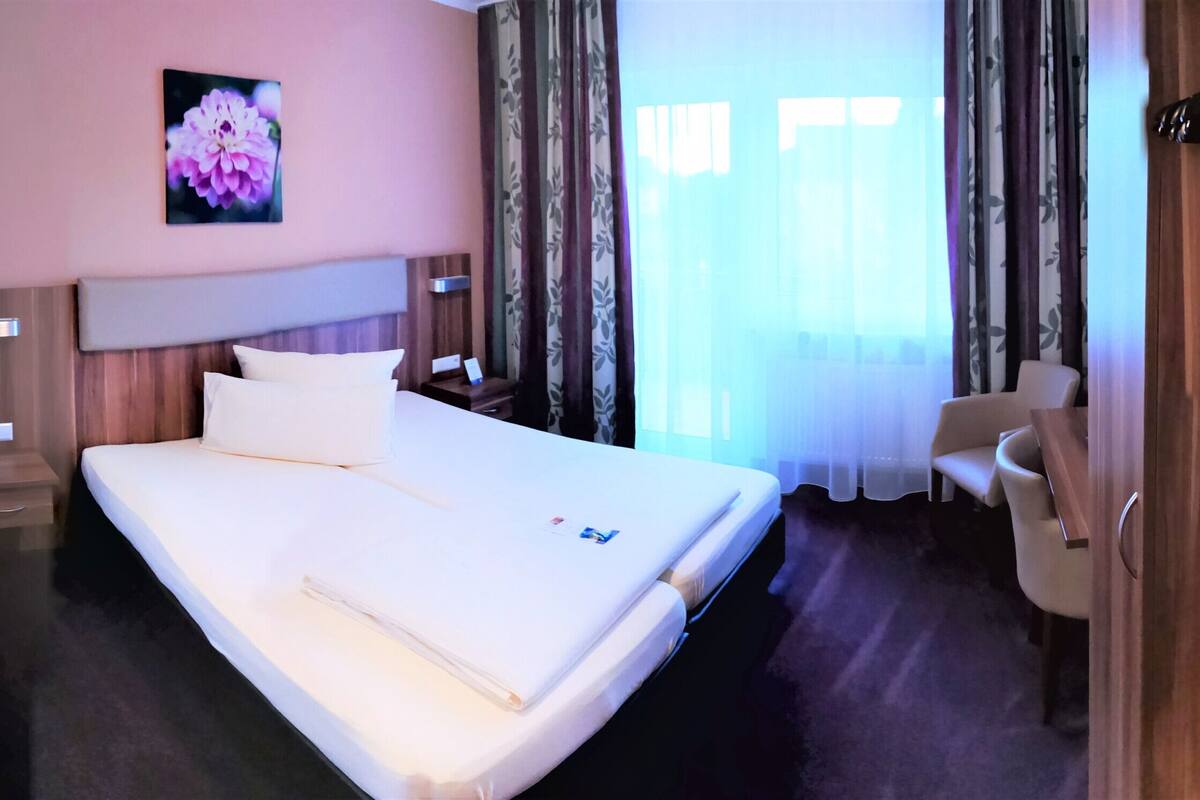A hotel room with a bed, a bedside table, a chair, a shower area, and a window with curtains.