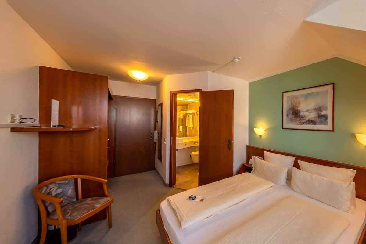 A hotel room with a bed, a chair, a desk, a shelf with toiletries, and a painting on the wall.
