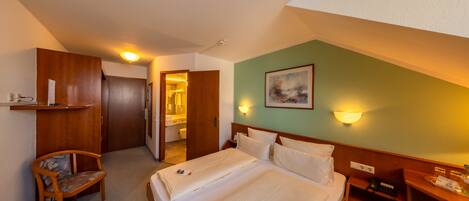 Una habitación de hotel con cama, una silla, un escritorio, repisas con artículos de aseo y un cuadro en la pared.