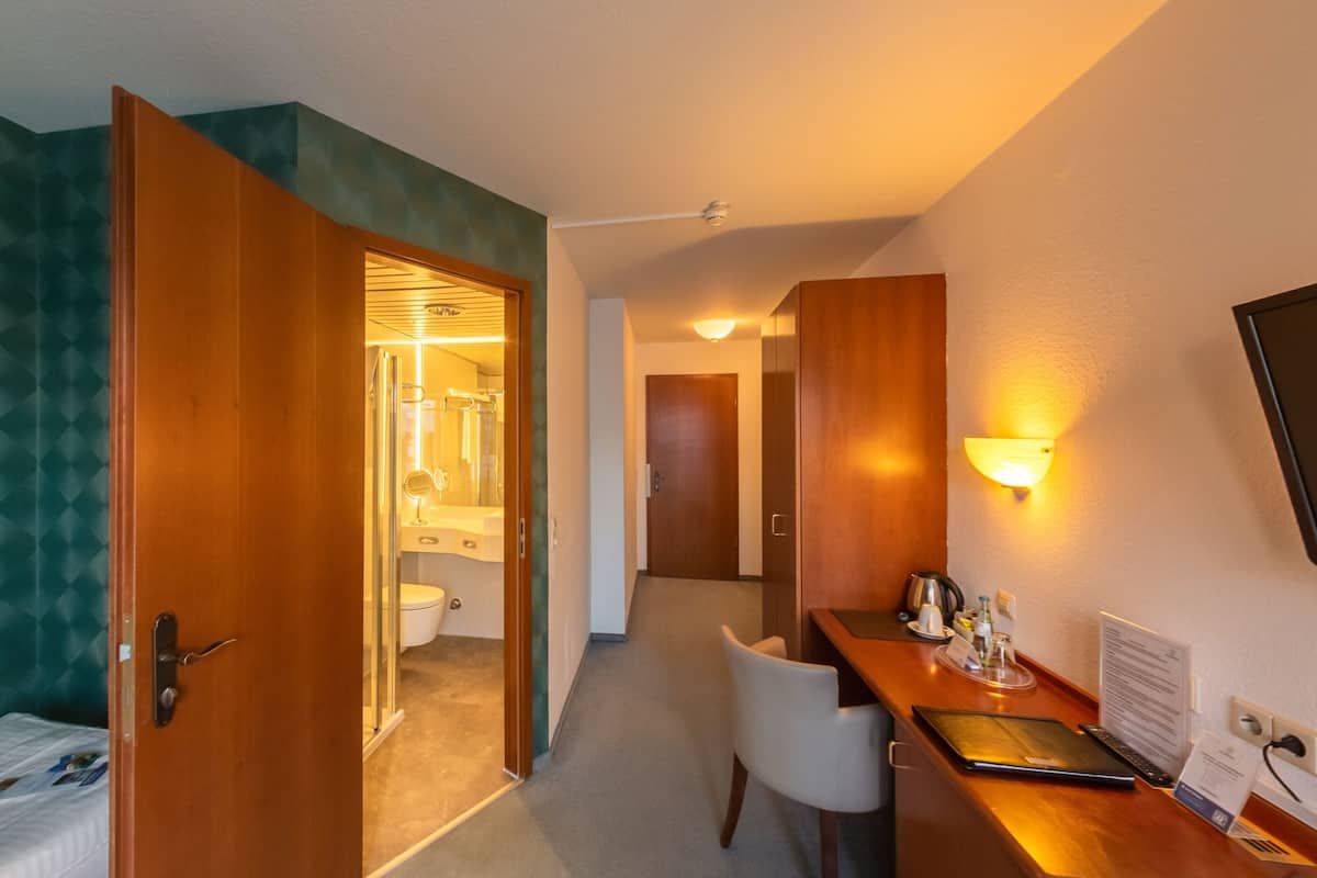A hotel room with a bed, a desk with a chair, a TV, and a bathroom visible through an open door.