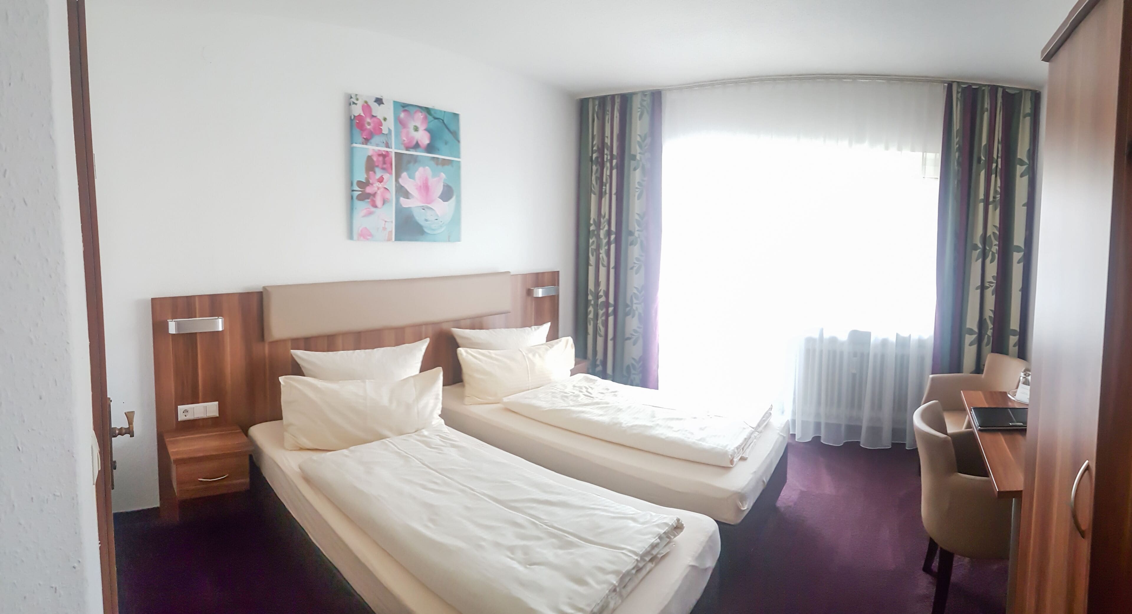 Habitación de hotel con dos camas, cabecero de madera, mesita de noche pequeña, escritorio con silla y ventana con cortinas.