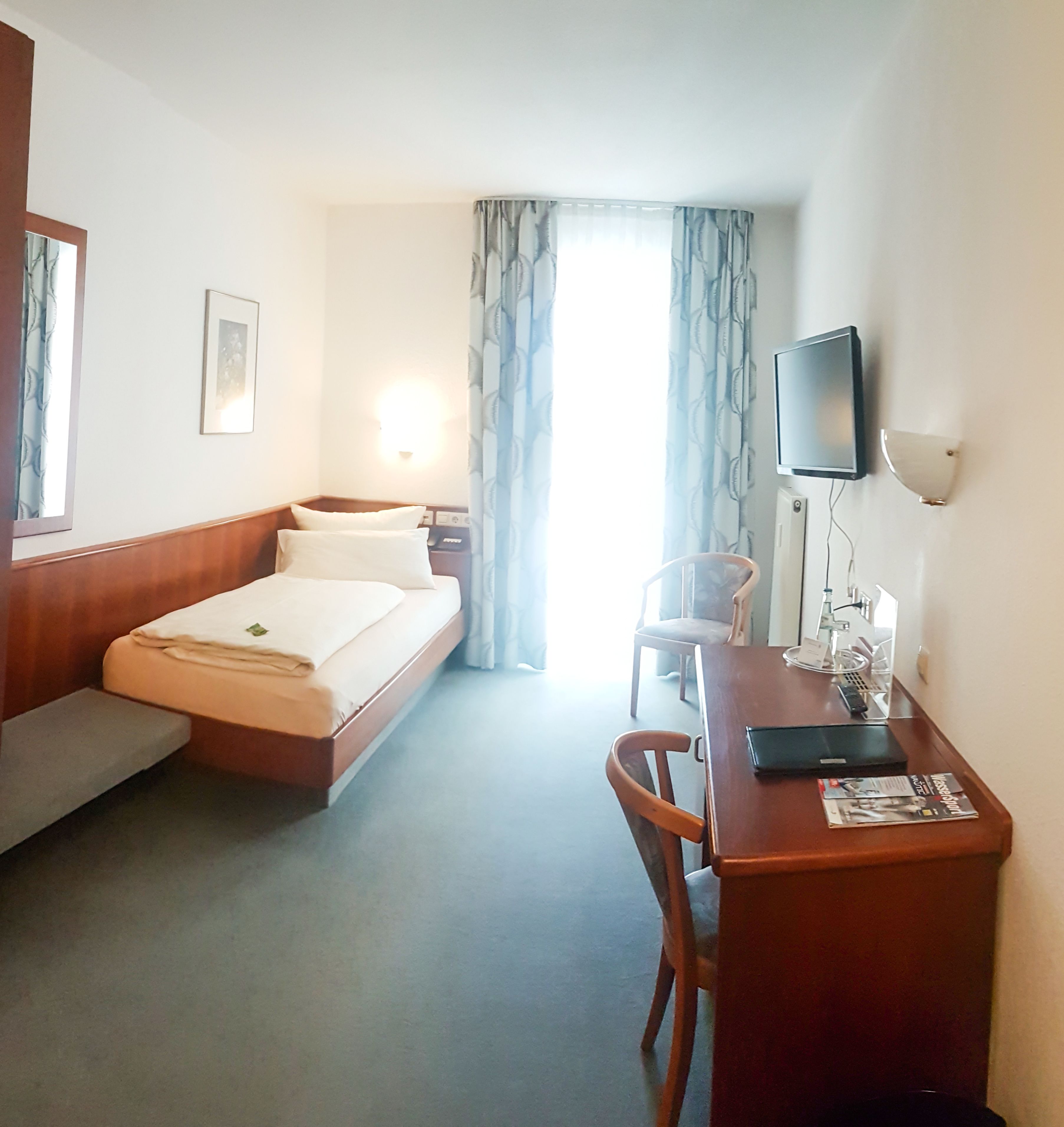 A hotel room with a bed, a desk with a chair, a TV, and a window with curtains.
